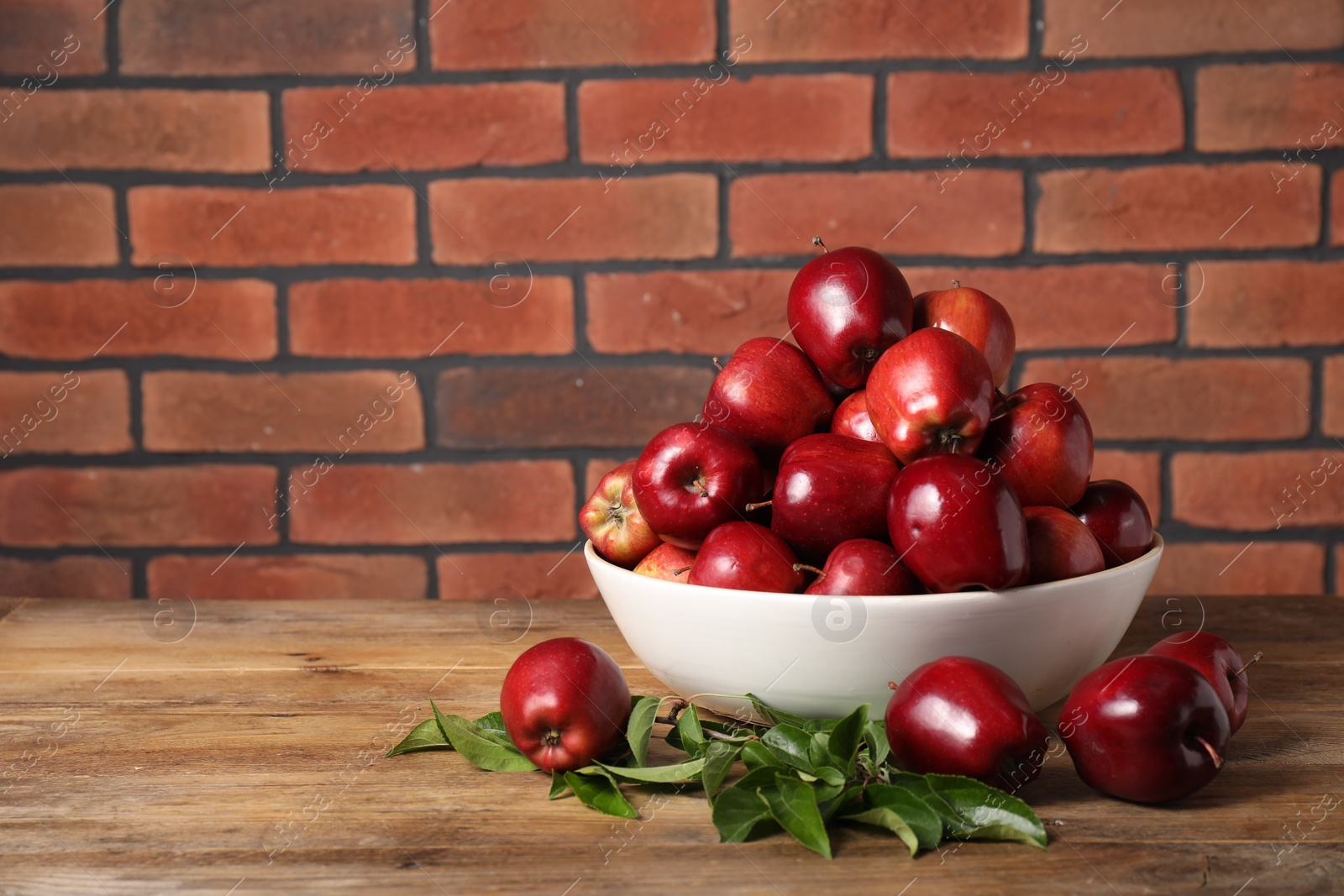 Photo of Fresh ripe red apples in bowl and leaves on wooden table. Space for text