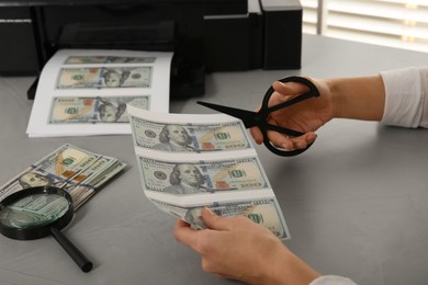 Photo of Counterfeiter cutting dollar banknotes with scissors at table indoors, closeup. Fake money concept