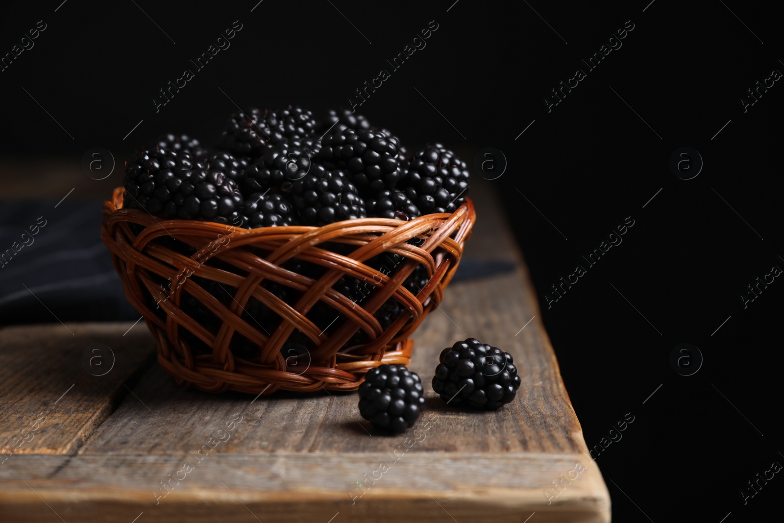 Photo of Fresh ripe blackberries in wicker bowl on wooden table against dark background
