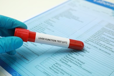 Laboratory worker holding tube with blood sample and label Liver Function Test near form at table, closeup. Space for text
