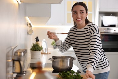 Photo of Smiling woman with wooden spoon tasting soup in kitchen