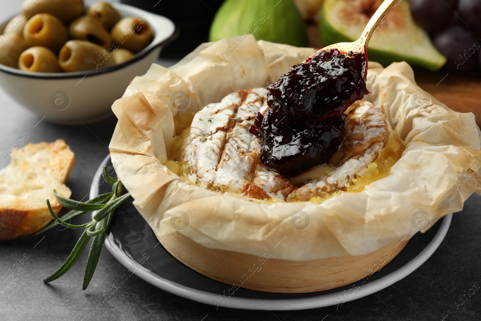 Photo of Putting jam onto tasty baked brie cheese on grey table, closeup