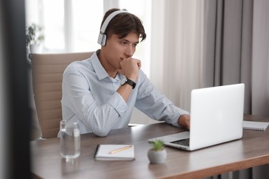 Photo of Man in headphones watching webinar at wooden table in office