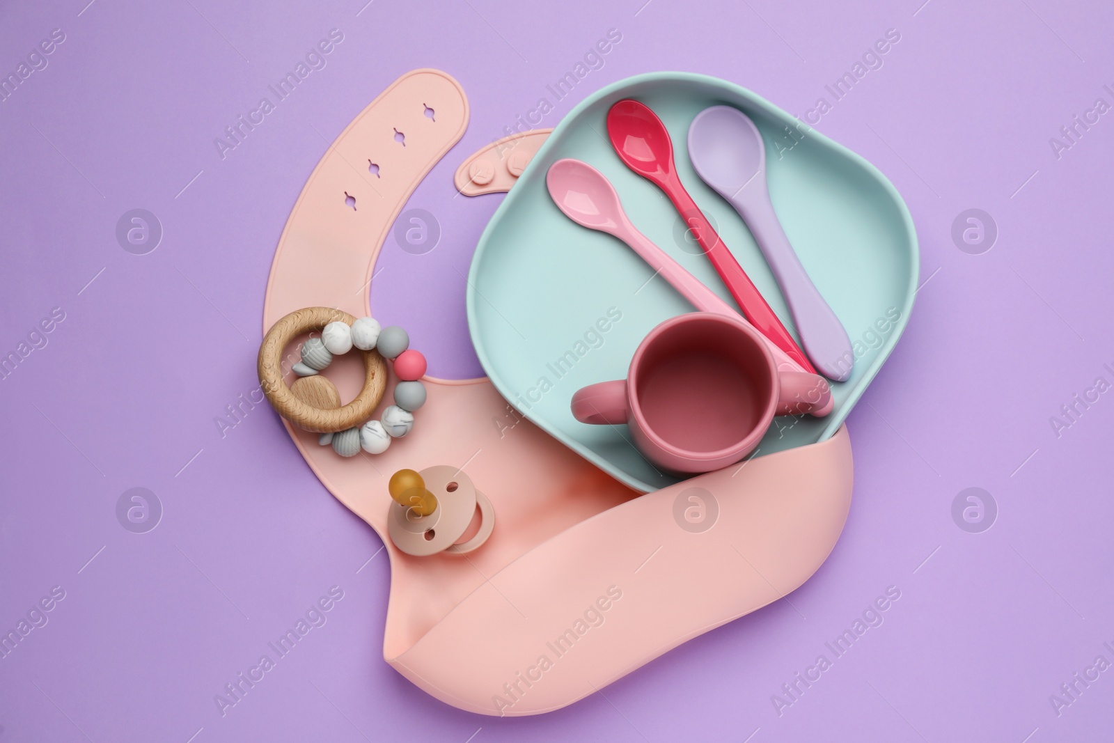 Photo of Set of plastic dishware, silicone bib and baby accessories on violet background, flat lay