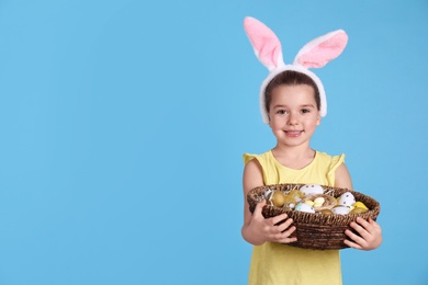 Happy little girl with bunny ears holding wicker basket full of Easter eggs on light blue background. Space for text