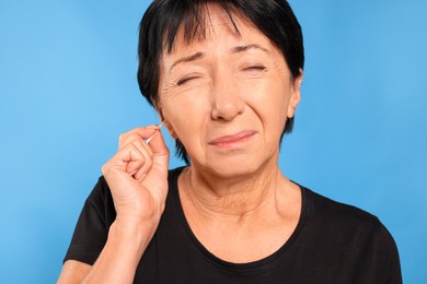 Senior woman cleaning ear with cotton swab on light blue background, closeup