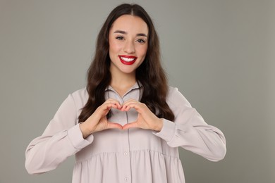 Beautiful young woman making heart with hands on grey background
