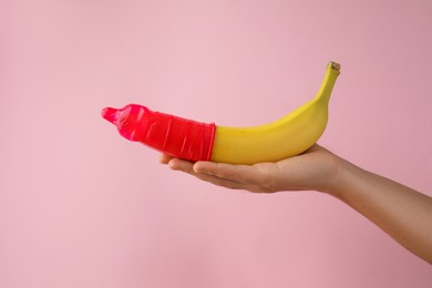 Photo of Woman holding banana in condom on pink background, closeup. Safe sex concept