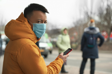 Photo of Asian man with smartphone wearing medical mask on city street, space for text. Virus outbreak