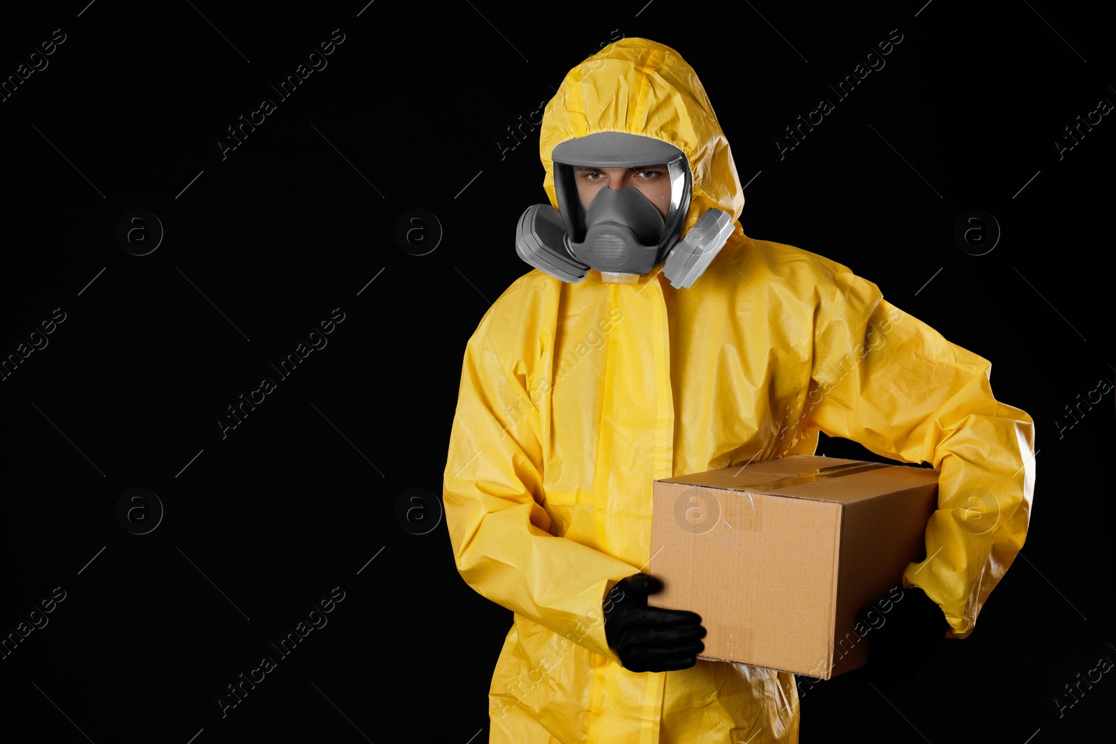 Photo of Man wearing chemical protective suit with cardboard box on black background, space for text. Prevention of virus spread