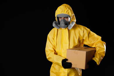 Photo of Man wearing chemical protective suit with cardboard box on black background, space for text. Prevention of virus spread