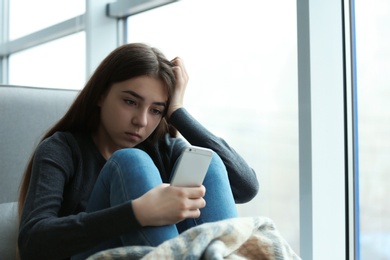 Upset teenage girl with smartphone sitting at window indoors. Space for text