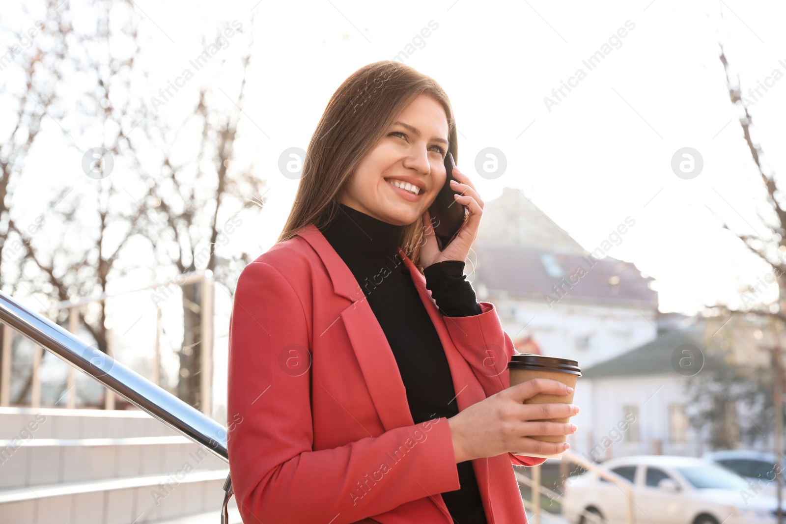 Photo of Businesswoman with cup of coffee talking on smartphone on city street in morning