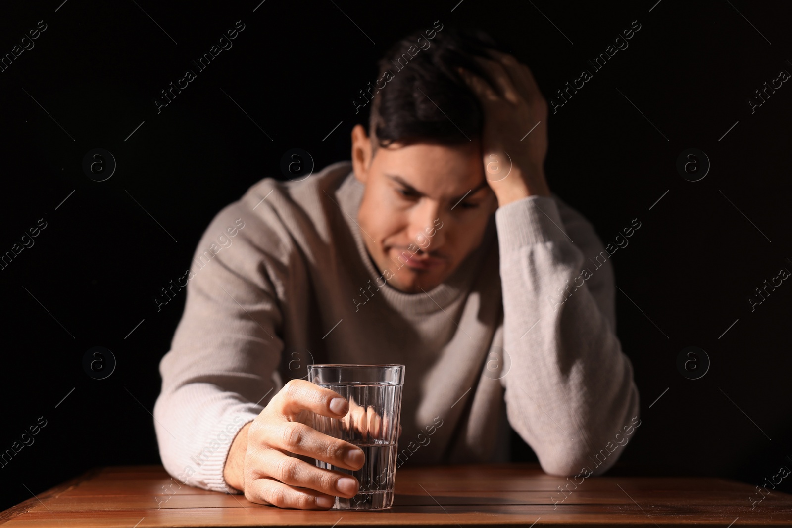 Photo of Addicted man at wooden table against black background, focus on glass of alcoholic drink