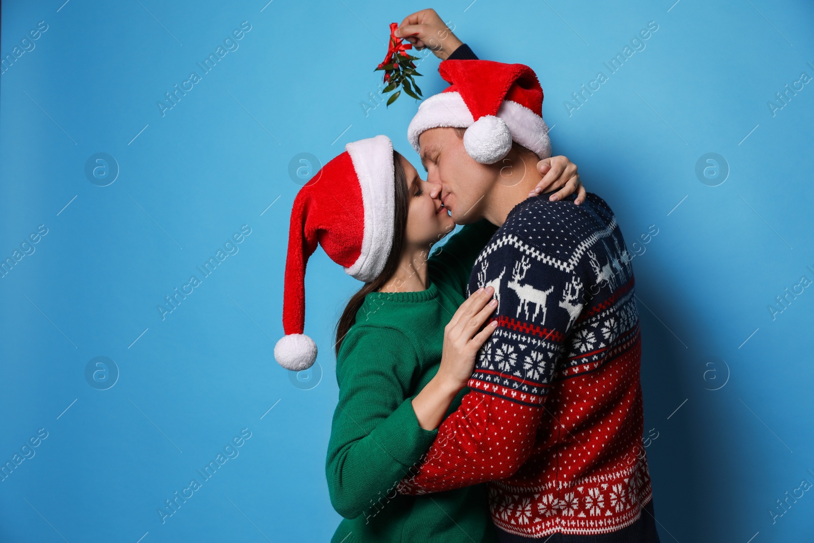 Photo of Happy couple kissing under mistletoe bunch on light blue background