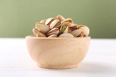 Photo of Tasty pistachios in bowl on white wooden table against olive background, closeup