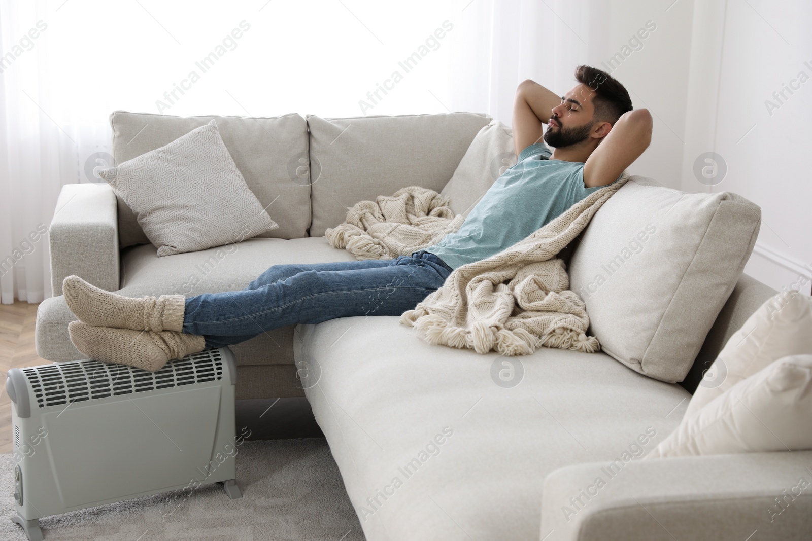 Photo of Young man warming feet on electric heater at home