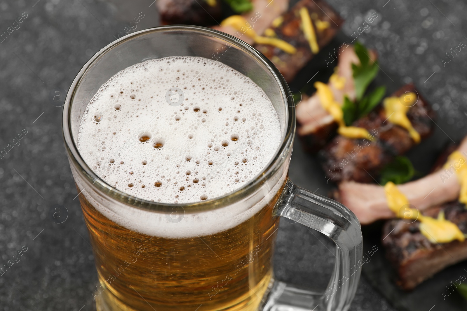 Photo of Mug with beer and delicious grilled ribs on dark grey textured table, closeup