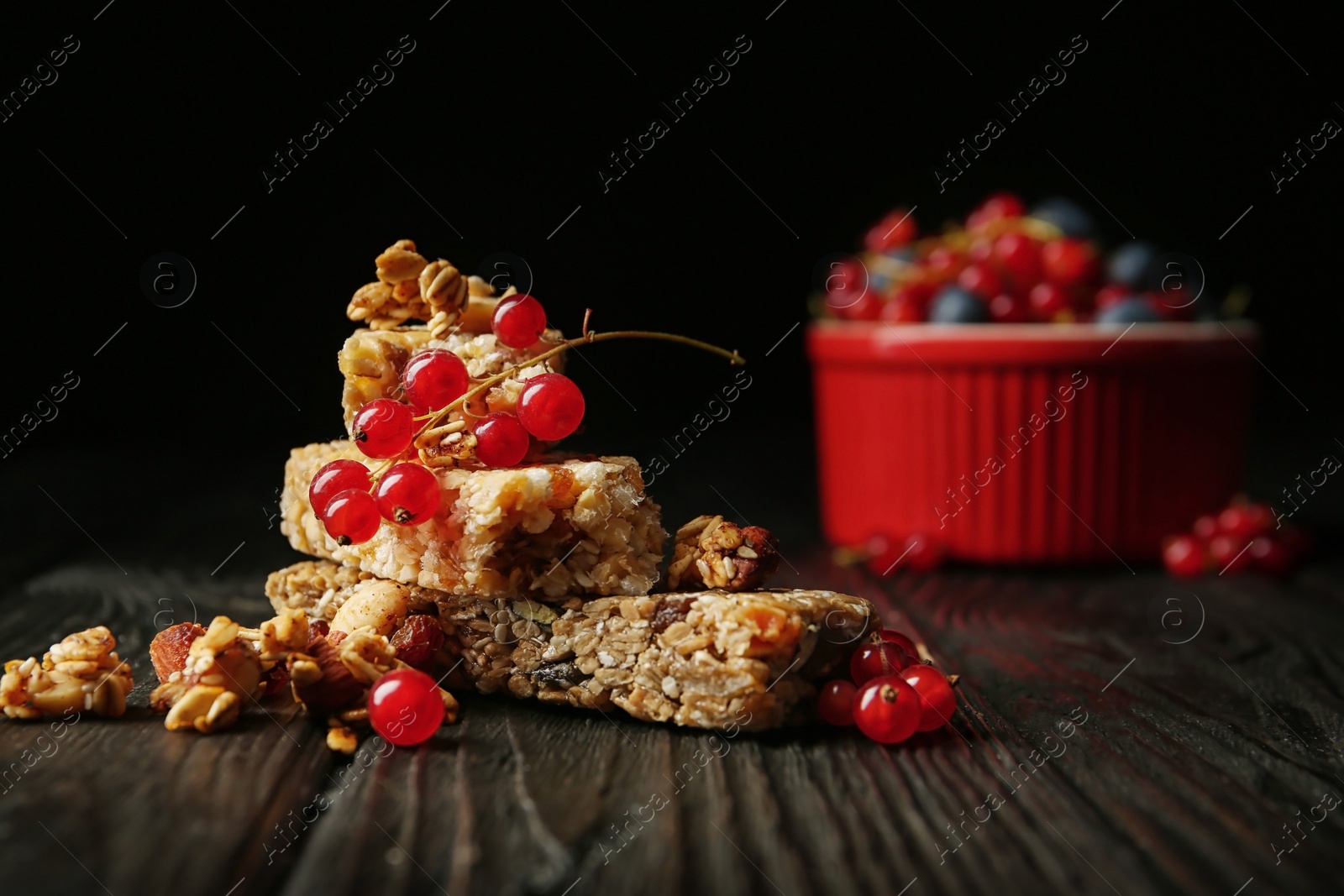 Photo of Different grain cereal bars and red currant on wooden table against black background