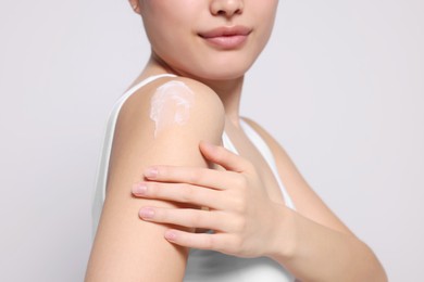 Young woman applying body cream onto shoulder on light grey background, closeup