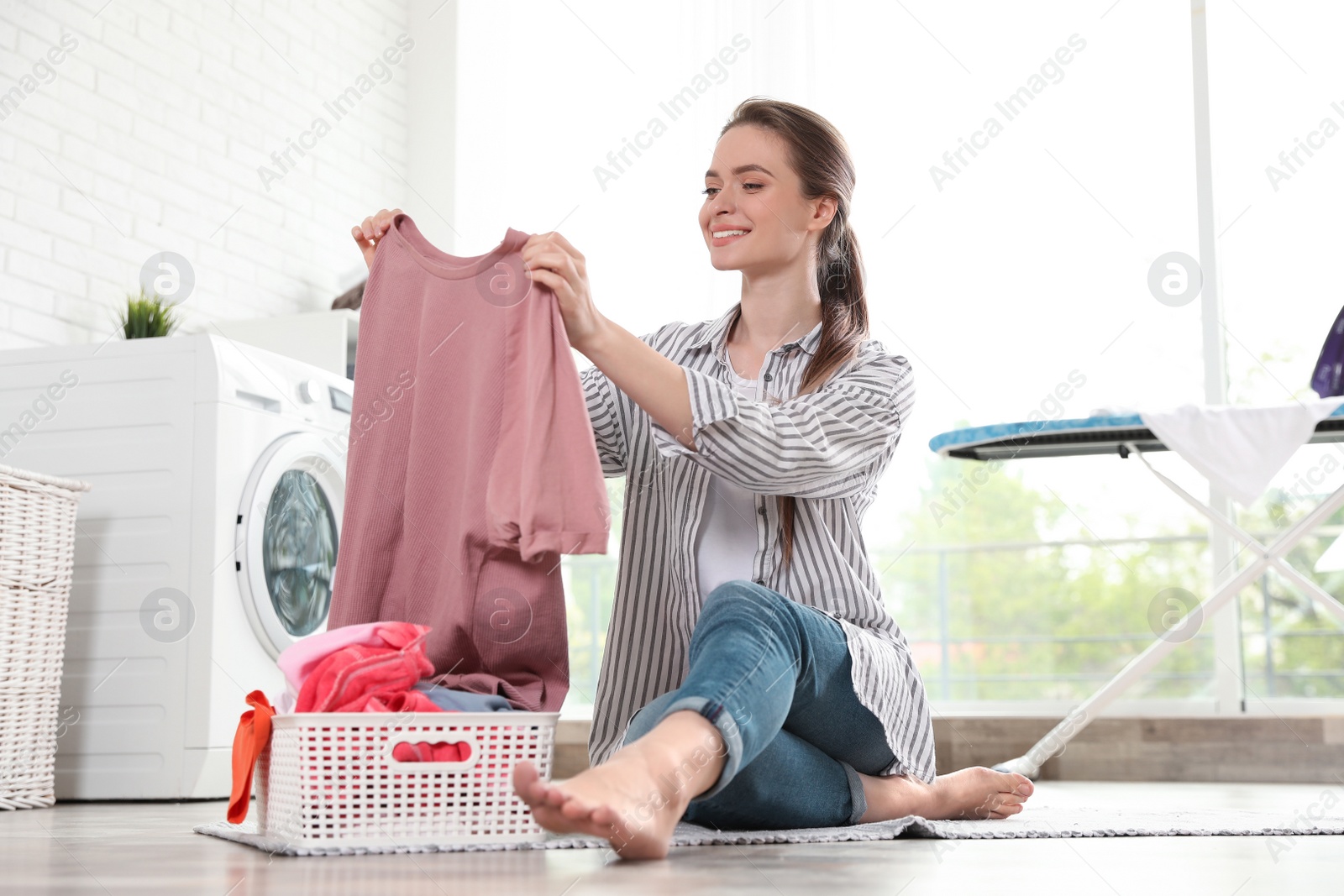 Photo of Young woman with basket of clean laundry on floor in room