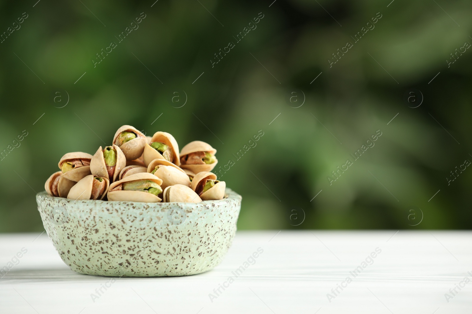 Photo of Tasty pistachios in bowl on white table against blurred background. Space for text