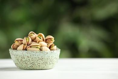 Tasty pistachios in bowl on white table against blurred background. Space for text