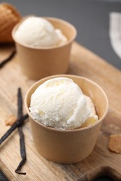 Paper cups with delicious ice cream and vanilla pods on wooden board, closeup