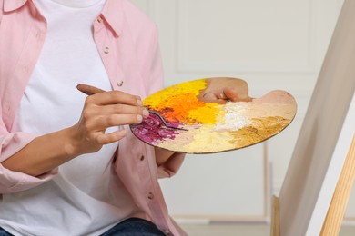 Photo of Woman mixing paints on palette near easel in studio, closeup