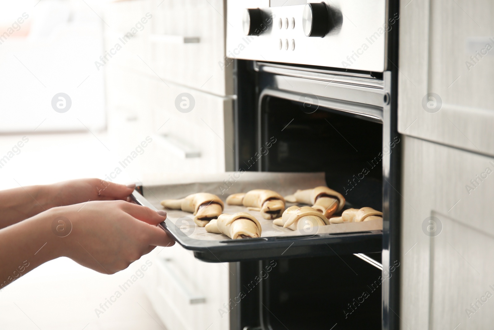 Photo of Woman putting baking sheet with raw croissants in oven, closeup