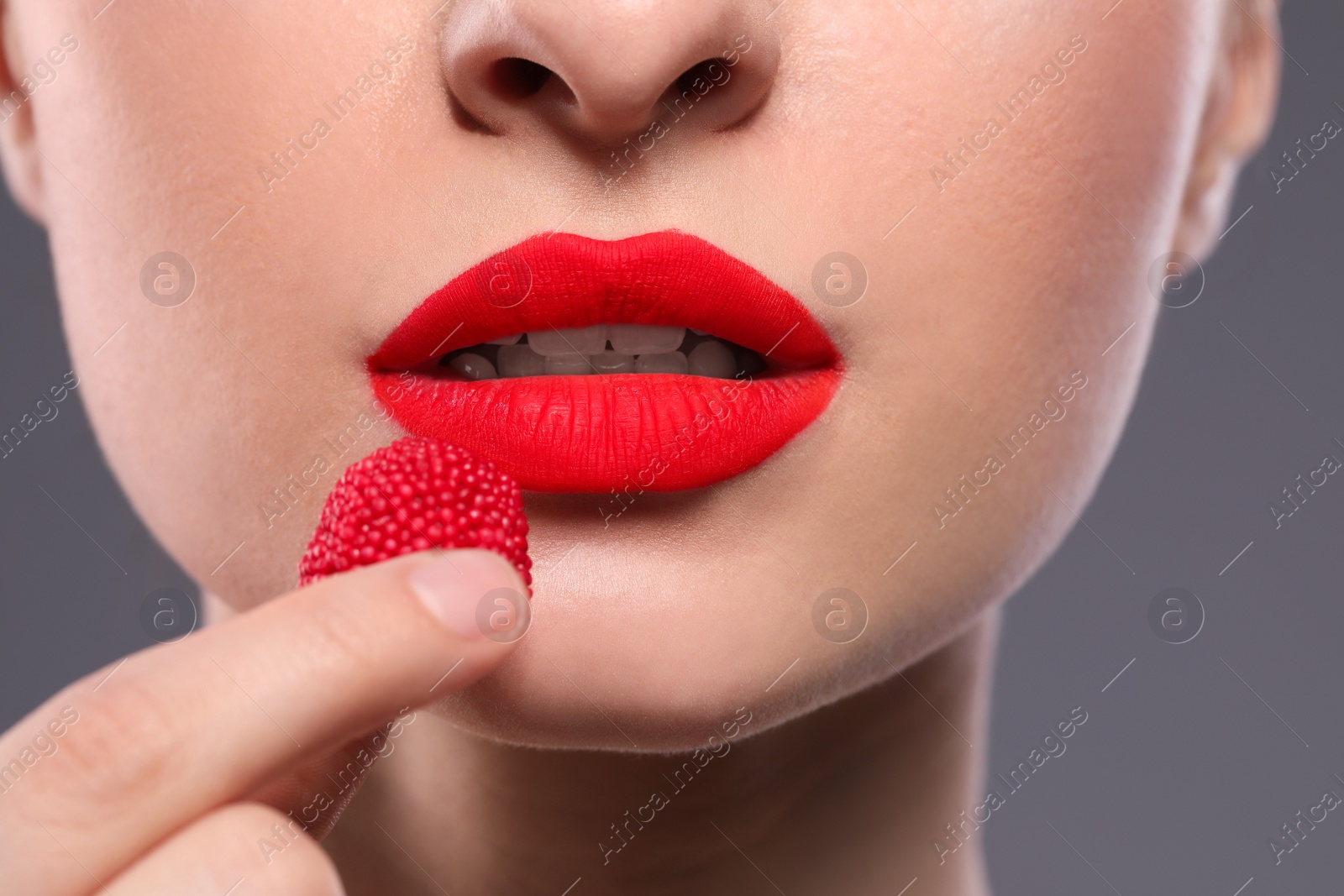 Photo of Young woman with beautiful red lips makeup eating candy on grey background, closeup