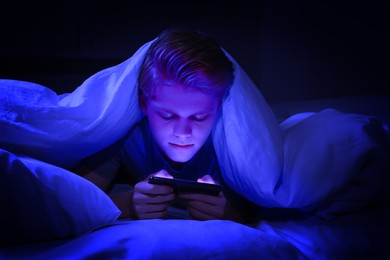 Image of Internet addiction. Teenage boy using smartphone under blanket on bed at night. Toned in blue