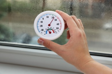 Photo of Woman holding round hygrometer with thermometer near window on rainy day, closeup