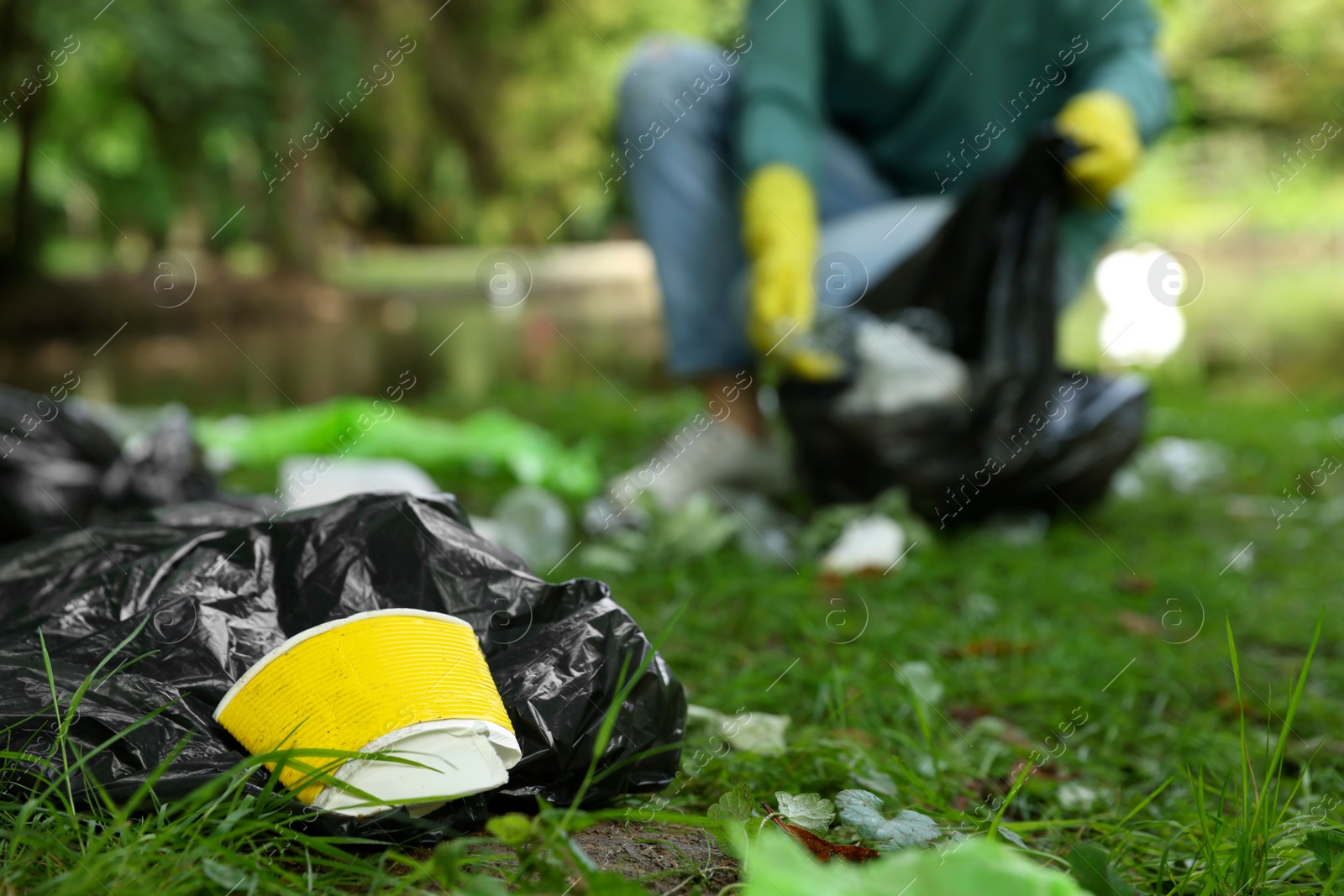 Photo of Woman with plastic bag collecting garbage in park, focus on paper cup