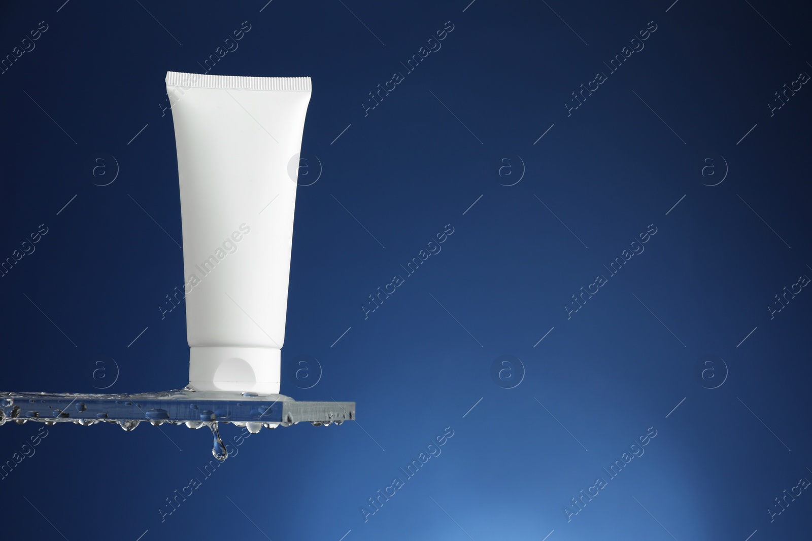 Photo of Moisturizing cream in tube on glass with water drops against blue background. Space for text