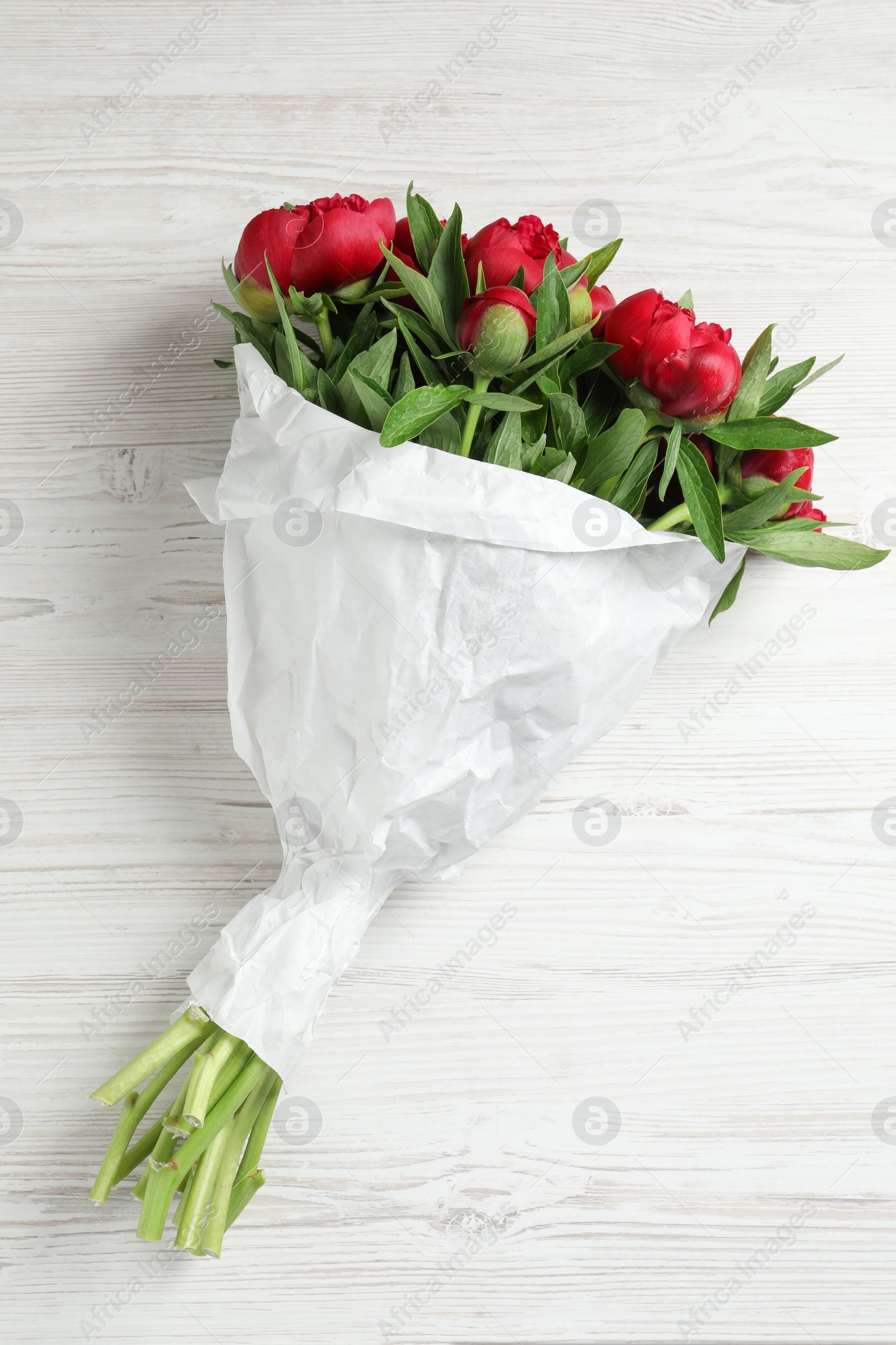 Photo of Beautiful bouquet of red peony flowers wrapped in paper on white wooden table, top view