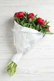 Beautiful bouquet of red peony flowers wrapped in paper on white wooden table, top view