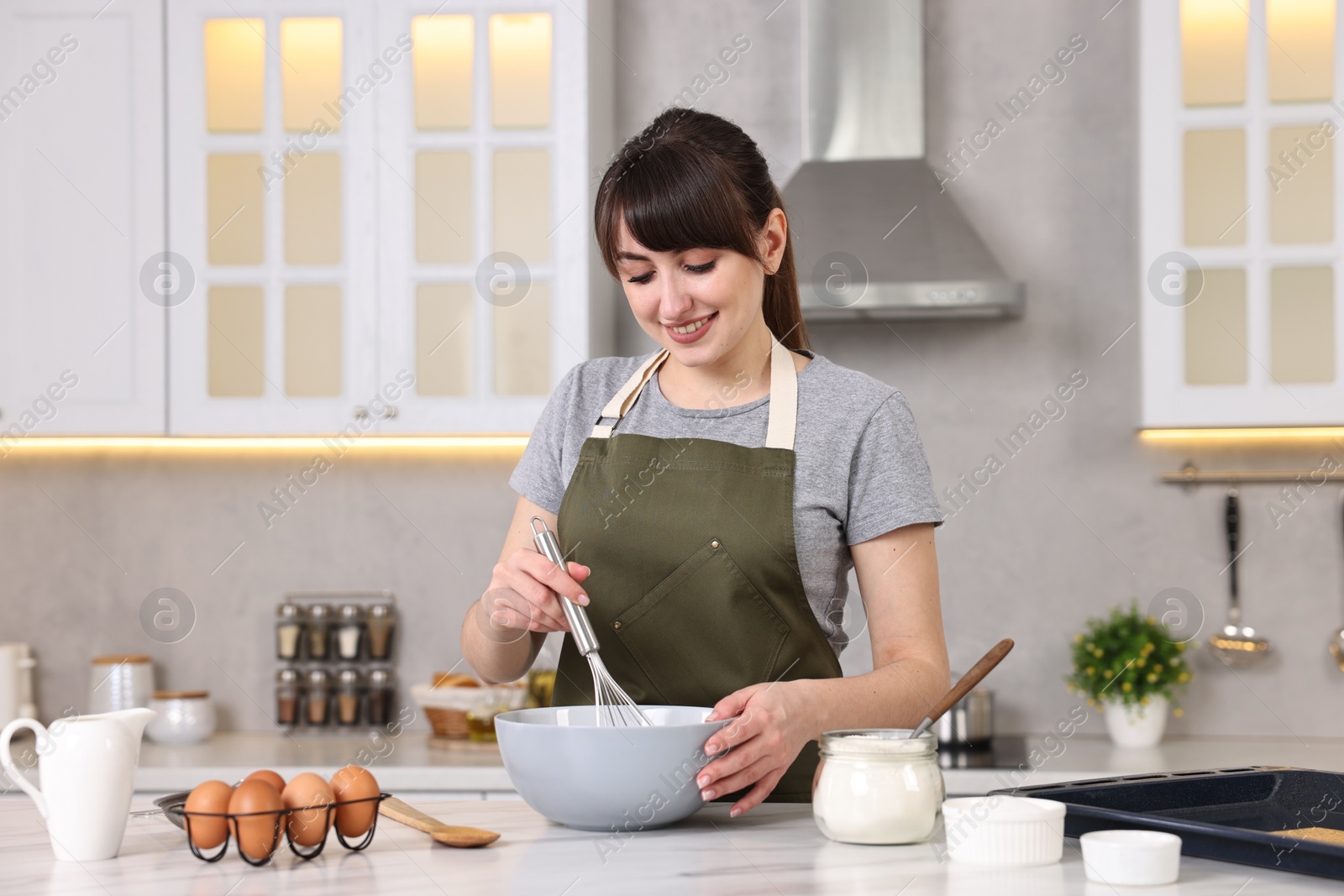 Photo of Happy young housewife mixing products into bowl at white marble table in kitchen