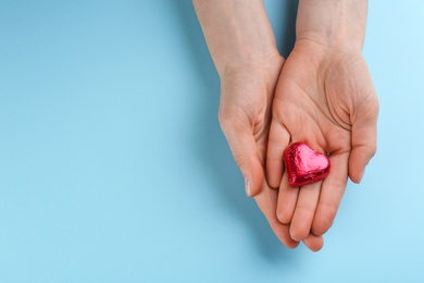 Woman holding heart shaped chocolate candy on light blue background, top view. Space for text