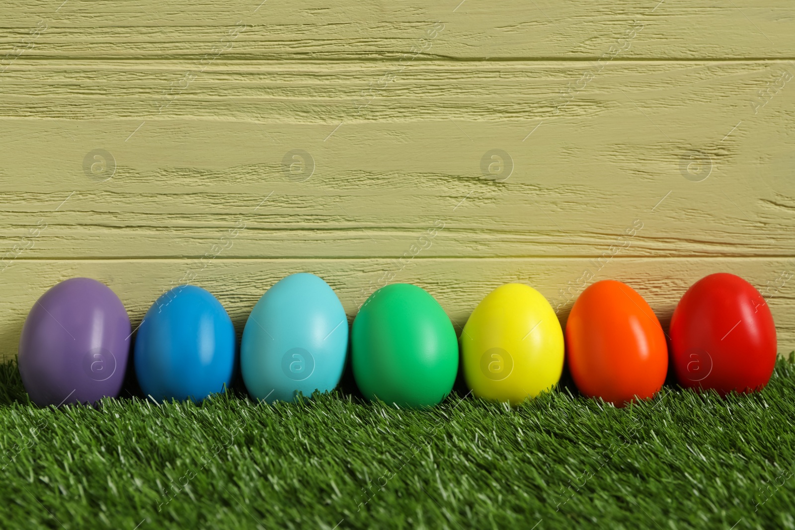 Photo of Bright Easter eggs on green grass against yellow wooden background