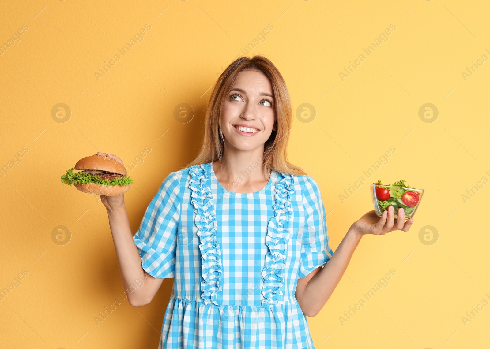 Photo of Young woman holding burger and salad on color background. Choice between diet and unhealthy food