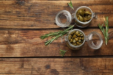 Photo of Tasty capers in jars and rosemary on wooden table, flat lay. Space for text