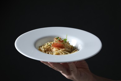 Woman holding plate of tasty spaghetti with prosciutto and microgreens on black background, closeup. Exquisite presentation of pasta dish