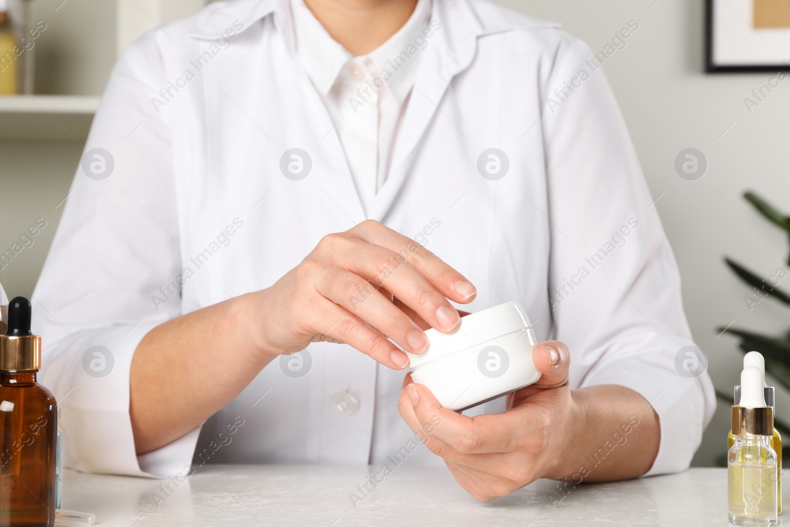 Photo of Dermatologist holding jar of cream at white table indoors, closeup. Developing cosmetic product