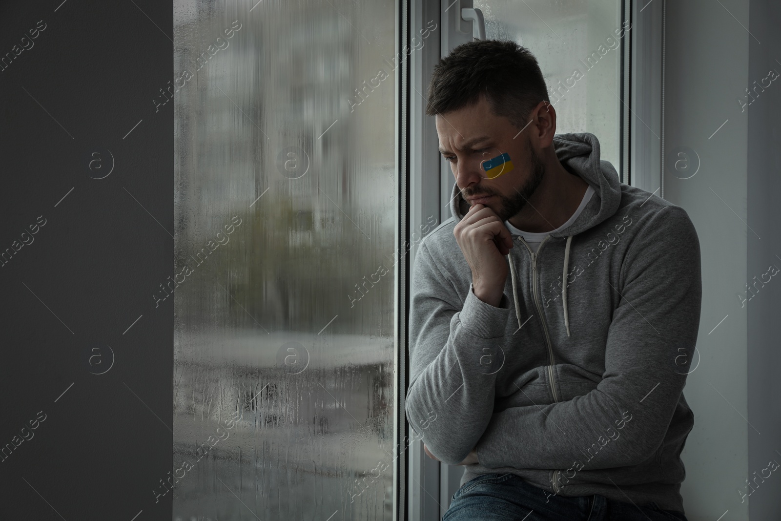 Image of Upset man with picture of Ukrainian flag on cheek near window at home, space for text. Stop war in Ukraine