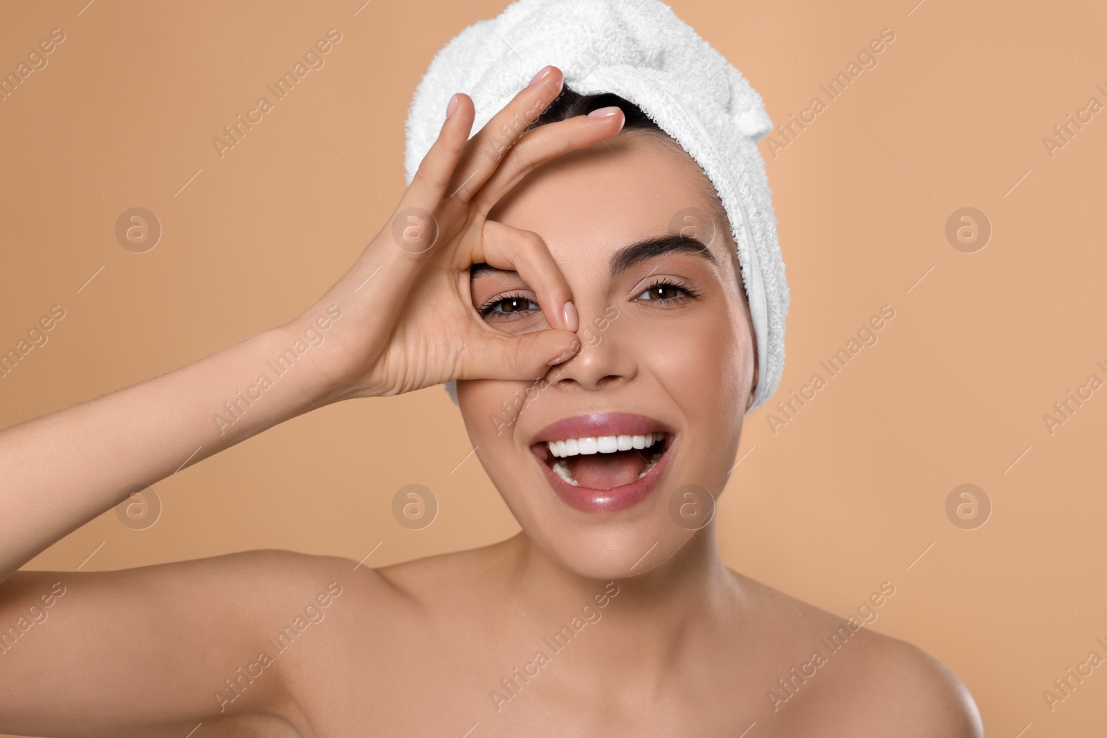 Photo of Woman with towel showing ok gesture near her eye on beige background. Spa treatment