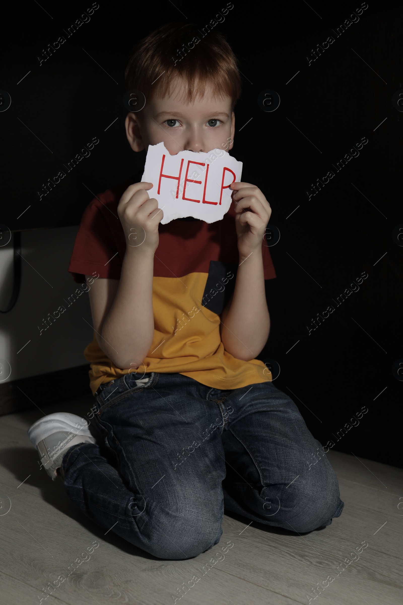 Photo of Scared little boy holding piece of paper with word Help under table on floor indoors. Domestic violence concept