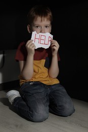 Photo of Scared little boy holding piecepaper with word Help under table on floor indoors. Domestic violence concept