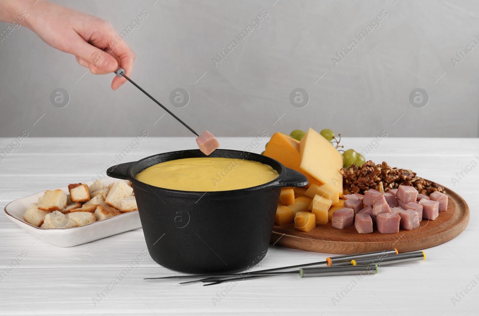 Photo of Woman dipping piece of ham into fondue pot with tasty melted cheese at white wooden table, closeup