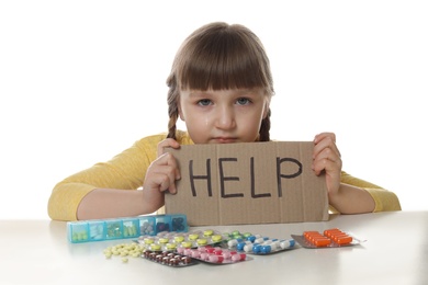 Photo of Crying little child with many different pills and word Help written on cardboard against white background. Danger of medicament intoxication